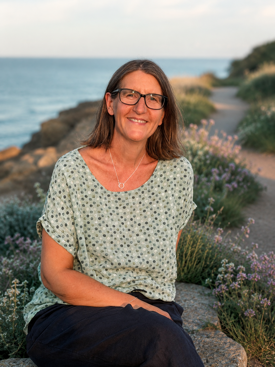 Anna Blair, cancer coach, sitting on a coastal path with the sea behind her